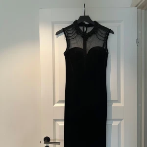 Svart långklänning med pärldetaljer - Säljer en svart långklänning med transparent mesh upptill och dekorativa svarta pärlor runt halsen och axlarna. Klänningen är ärmlös och har en elegant, figurnära passform. Perfekt för fest tillfällen! Storleken är S och klänningen är köpt i London! Den saknar några pärlor på vissa ställen men är man kreativ kan man säkert brodera tillbaka liknande pärlor eller komma på något annat kreativt, eller så kan man ha på sig den som den är 🥰