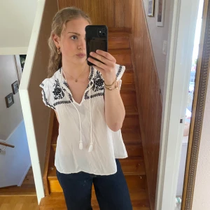 Vit blus med broderade detaljer - Vit blus med svarta broderade mönster vid axlar och halsringning. Blusen har korta ärmar med volangkant och knytband framtill. Luftig och lätt i materialet, perfekt för varmare dagar. Blusen är aldrig använd
