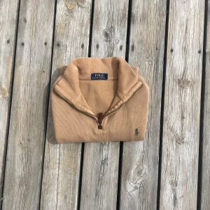 Beige stickad tröja med halv dragkedja från Polo Ralph Lauren - Säljer en beige stickad tröja från Polo Ralph Lauren med halv dragkedja och broderad logga på bröstet. Tröjan har hög krage och är perfekt för lager-på-lager. Klassisk och stilren design.