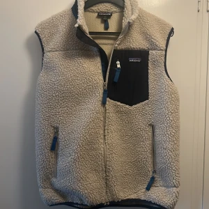Beige fleeceväst från Patagonia - Säljer en beige fleeceväst från Patagonia med dragkedja framtill och svart bröstficka med dragkedja. Västen har blå detaljer på dragkedjorna och är perfekt för lager-på-lager. Klassisk och tidlös design.