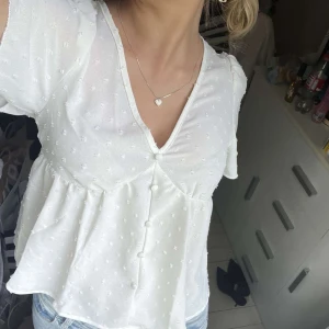 Vit blus med knappar och v-ringning - Söt vit blus med v-ringning och dekorativa knappar framtill. Blusen har korta ärmar och ett diskret prickigt mönster i tyget. Lätt och luftig modell som passar perfekt till jeans. (Oanvänd)