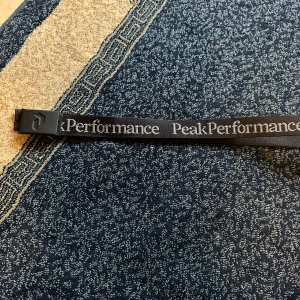 Svart bälte från Peak Performance - Säljer ett svart bälte från Peak Performance med tydlig logga längs hela bandet och en robust svart spänne. Bältet är i textilmaterial och passar perfekt till jeans eller andra byxor. Snygg och stilren accessoar för att lyfta din outfit.