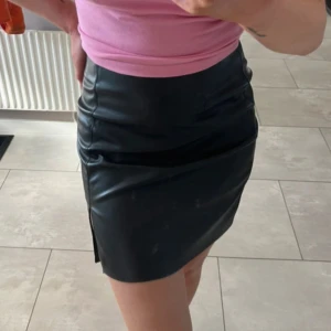 Svart skinnkjol med slits - Säljer en  skinnkjol med tight passform och liten slits på sidan. Kjolen är kort och har en clean, stilren look som passar perfekt till en snygg topp eller t-shirt.