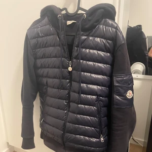 Mörkblå hybridjacka med huva från Moncler - Snygg mörkblå jacka från Moncler med quiltad framsida och ärmar i sweatshirtmaterial. Jackan har huva, dragkedja och Moncler-logga på ärmen. Perfekt för lager-på-lager och streetwear-stil.