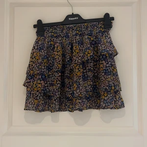 Blommig volangkjol från KappAh - Supersöt kort kjol med blommigt mönster i blått, gult och vitt från KappAhl. Kjolen har flera volanger och en luftig känsla, perfekt för varmare dagar.barnstorlek men passar xxs och xs