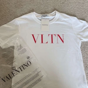 Vit t-shirt med röd VLTN-logga från Valentino - Snygg vit t-shirt från Valentino med stor röd VLTN-logga på bröstet. Klassisk rund halsringning och korta ärmar. Tillverkad i mjuk bomull och perfekt för en clean och trendig look. Kvitto finns också
