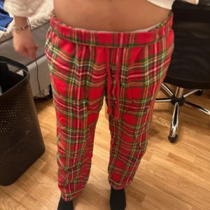 Röda rutiga mjukisbyxor med snörning - Säljer ett par röda mjukisbyxor med grönt och gult rutmönster. Byxorna har resår i midjan och snörning framtill för extra komfort. Perfekta för att chilla hemma eller mysa runt i.