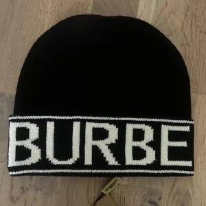 Svart mössa från Burberry - Svart mössa från Burberry med bred, vit logotyp runt kanten. Klassisk och stilren design med uppvikt kant. Perfekt accessoar för att hålla värmen och samtidigt addera en lyxig touch till din outfit.