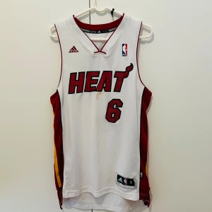 Miami Heat baskettröja LeBron James #6 - Säljer en vit Miami Heat baskettröja från Adidas med LeBron James och nummer 6. Tröjan har röda och gula detaljer på sidorna, broderade loggor och är ärmlös. Perfekt för basketfans som vill sticka ut på planen eller på läktaren.