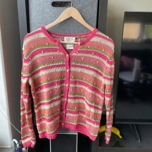 Randig kofta från Beyond retro - Sönder på baksidan! Säljer en färgglad stickad kofta från Beyond retro i rosa, beige, vitt och orange. Koftan har långa ärmar, knappar framtill och ett randigt mönster med små dekorativa pärlor. Perfekt för dig som gillar unika detaljer och mjuka material.