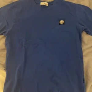 Blå t-shirt från Stone Island - Säljer en blå t-shirt från Stone Island med klassisk logotyp-patch på bröstet. T-shirten har rund hals och korta ärmar. Perfekt för dig som gillar stilrena plagg med ikoniska detaljer. Sedan finns det ett håll på sidan som går att sy ihop rätt så lätt. ABS! Pris går att diskutera.