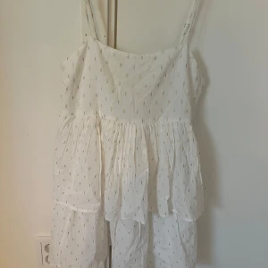 Vit sommarklänning med tunna axelband - Söt vit klänning med tunna axelband och små broderade detaljer i guld. Klänningen har en luftig, volangig kjol och smock i ryggen för extra komfort. Perfekt för varma dagar och enkel att styla med sandaler eller sneakers.