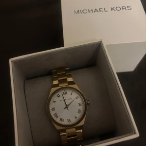 Guldfärgad klocka från Michael Kors - Stilren guldfärgad klocka från Michael Kors med vit urtavla och romerska siffror. Klockan levereras i originalask med extra länk och manual. Perfekt accessoar för att lyfta din outfit. Pris kan diskuteras vid snabb affär 