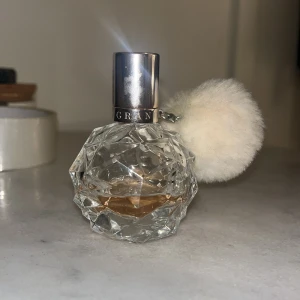 Ariana Grande parfym med fluffig vit boll - Säljer en parfym från Ariana Grande i en kristallformad, transparent flaska med silverfärgad kork och en stor fluffig vit boll som dekoration. Flaskan har ett lyxigt och unikt utseende, perfekt för dig som gillar snygga detaljer på sminkbordet.