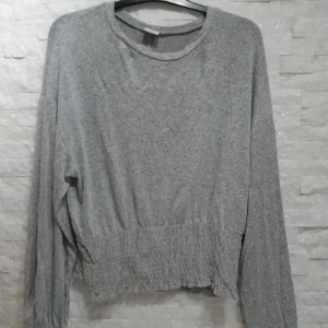 Grå långärmad tröja med ribbad midja - Mysig grå tröja med lång ärm och bred ribbad kant nedtill. Tröjan har en avslappnad passform och är tillverkad i ett mjukt, stickat material. Perfekt för en chill och stilren look.