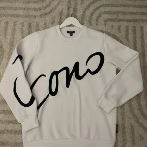 Vit sweatshirt från Icono med svart text - Säljer en vit sweatshirt från Icono med stor svart text över framsidan. Tröjan har rund halsringning och långa ärmar. Perfekt för en stilren och avslappnad look.