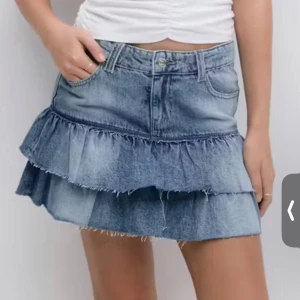 Jeanskjol med volang - Snygg blå jeanskjol med volangdetaljer och rå kant nertill. Kjolen har klassisk femficksdesign och stängs med knapp och dragkedja framtill. Den är helt oanvänd, ny utan prislapp. Perfekt för dig som gillar en trendig och avslappnad stil.