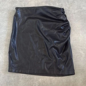 Svart kort kjol i skinnimitation - Säljer en svart kort kjol i skinnimitation med snygg rynkdetalj på ena sidan och dragkedja på andra sidan. Kjolen har en tight passform och är perfekt för dig som gillar en edgy stil. Jätte fin och stilig.