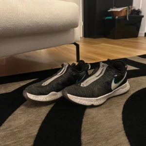 Svarta Nike sneakers med dragkedja - Säljer ett par svarta Nike sneakers med vit sula och dragkedja framtill. Skorna har diskret mönster i vitt och grått samt en blå Nike-logga på sidan. Perfekta för dig som gillar sportig stil och vill ha något unikt.