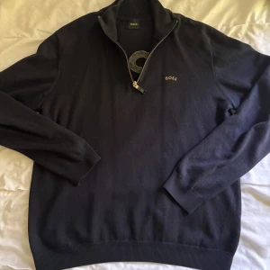 Svart tröja med dragkedja från BOSS - Säljer en svart långärmad tröja från BOSS med halv dragkedja och broderad logga på bröstet. Tröjan har ribbade muddar och en stor BOSS-logga på ryggen i gult. Perfekt för en stilren och avslappnad look.