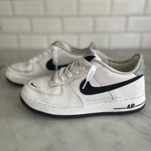 Nike Air Force 1 vita sneakers med svart swoosh - Säljer ett par klassiska Nike Air Force 1 sneakers i vitt med svart swoosh och grå detalj på hälen. Skorna har snörning, rund tå och platt sula. Perfekta för dig som gillar en tidlös och sportig stil. Storlek 40 EU, bra skick men använda