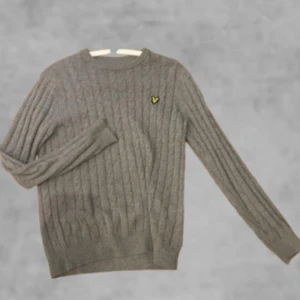 Grå kabelstickad tröja från Lyle & Scott - Säljer en klassisk grå kabelstickad tröja från Lyle & Scott med rund halsringning och den ikoniska gula loggan på bröstet. Tröjan är mjuk och passar perfekt till lager-på-lager under kyliga dagar.