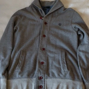 Grå cardigan från Fred Perry - Säljer en grå cardigan tröja från Fred Perry med knappar framtill och broderad logga på bröstet. Tröjan har långa ärmar, två fickor och snygga detaljer som synliga sömmar och bruna knappar. Perfekt för lager-på-lager-stil. Cardiganenr riktigt maskulin och kommer ytra din manlighet extra mycket. I jättebra skick. (Nyskick)