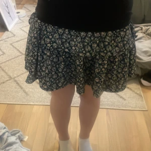 Blommig kortkjol i blått och vitt - Supersöt kortkjol med blommigt mönster i vitt och ljusrosa på mörkblå botten. Kjolen har en luftig och lätt form som ger en skön känsla. Perfekt för dig som gillar en feminin och avslappnad stil.