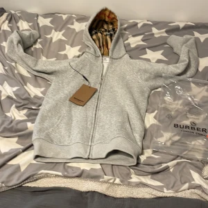 Grå hoodie från Burberry med dragkedja - Säljer en grå hoodie från Burberry med dragkedja och klassiskt rutigt foder i huvan. Tröjan har långa ärmar och ribbade muddar. Perfekt för en stilren och bekväm look. 