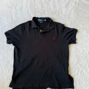Svart pikétröja från Polo Ralph Lauren M - Klassisk svart pikétröja från Polo Ralph Lauren med röd broderad logga på bröstet. Tröjan har korta ärmar, krage och knappar vid halsen. Piken är i skönt t shirt material 😊