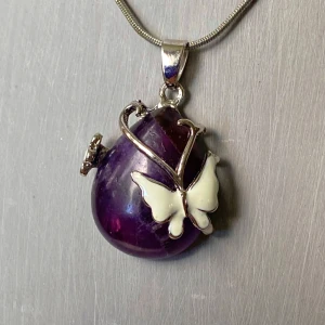 Lila halsband med fjärilshänge - Säljer ett halsband med ett droppformat lila hänge i ametist, dekorerat med silvriga detaljer och en vit fjäril framtill. Hänget sitter på en tunn silverfärgad kedja. Perfekt för dig som gillar unika smycken med naturinspiration.