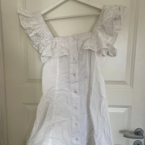 Vit klänning med volangärmar från Reclaimed Vintage - Superfin vit klänning från Reclaimed Vintage köpt från ASOS med breda volangärmar i broderad spets och knyt i ryggen. Klänningen har knappar framtill och en öppen ryggdetalj. Aldrig använd endast testad