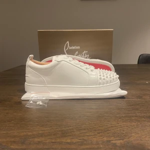 Vita sneakers från Christian Louboutin - Säljer ett par vita sneakers från Christian Louboutin med ikonisk röd sula och coola nitar framtill. Skorna har snörning och är tillverkade i skinn. Kommer med originalpåse och extra nitar. Perfekta för dig som vill sticka ut!