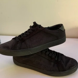 Svarta sneakers från Saint Laurent - Säljer ett par helsvarta sneakers från Saint Laurent med diskret logga på sidan och bak på hälen. Skorna har snörning, rund tå och är tillverkade i textil med detaljer i syntet vid hälen. Perfekta för en stilren look. Skick 8.5/10, storlek 39/40.