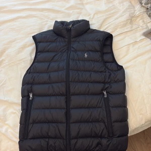 Svart dunväst från Polo Ralph Lauren - Säljer en svart dunväst från Polo Ralph Lauren med klassisk broderad logga på bröstet. Västen har hög krage, dragkedja framtill och två fickor med dragkedja. Perfekt för lager-på-lager och stilren look.