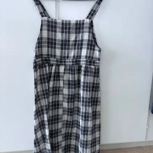 Rutig långklänning med knytband - Säljer en svartvit rutig långklänning med breda axelband och knytband i midjan. Klänningen har en rak passform och är tillverkad i ett mjukt bomullsliknande material. Perfekt för en avslappnad stil.