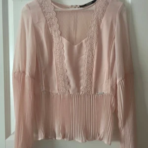 Ljusrosa blus med spets från Guess - Säljer en ljusrosa blus från Guess med långa ärmar, vacker spetsdetalj längs ringningen och plisserad nederdel. Blusen har en feminin och romantisk stil med fina detaljer framtill. Aldrig använd.