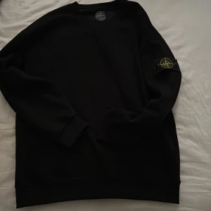 Svart sweatshirt från Stone Island - Säljer en svart sweatshirt från Stone Island med klassisk logotyp-patch på vänster ärm. Tröjan har rund halsringning och ribbade muddar vid ärmslut och nederkant. Perfekt för en stilren och avslappnad look.