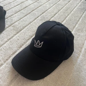Svart keps med vit krona - Svart keps med böjd skärm och justerbart spänne bak. Framtill finns en broderad vit krona. Perfekt accessoar för en stilren look.