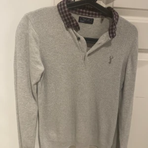 Grå tröja med rutigt krage från Next - Säljer en ljusgrå långärmad tröja från Next med stickad struktur upptill och rutigt krage i vinrött och grått. Tröjan har knappar vid halsen och en liten broderad detalj på bröstet. Perfekt för en stilren look. Priser är inte spikad i sten 