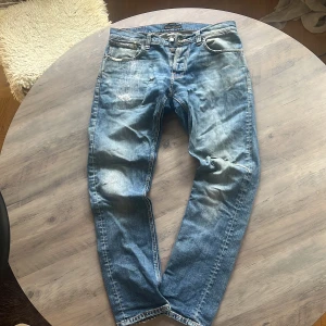 Blå jeans från Nudie Jeans - Säljer ett par klassiska blå jeans från Nudie Jeans med orange sömmar och knappar i gylfen. Jeansen har en normal passform och tydliga slitningar för en cool, använd look. Perfekta för dig som gillar en avslappnad stil. Nypris 2000kr mitt pris 449kr. Hör av er vid funderingar!🙌🎉