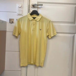 Gul piké t-shirt från Gant - Snygg gul piké t-shirt från Gant med klassisk krage och knappar framtill. Liten broderad logga på bröstet. Perfekt för en stilren och avslappnad look.