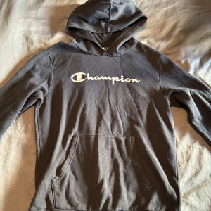 Svart hoodie från Champion - Mysig grå hoodie från Champion med vit logga över bröstet. Klassisk huva och stor magficka framtill. Perfekt för en avslappnad stil.