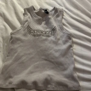 Vit ribbad linnetopp från Divided - Säljer en grå ribbad linnetopp från Cörkat med tryckt logga och vingar framtill. Toppen är ärmlös och har rund halsringning. Perfekt för en avslappnad och cool stil.