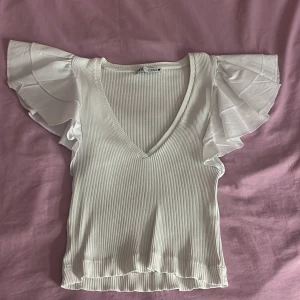 Vit ribbad topp med volangärmar från Zara - Supersöt vit ribbad topp från Zara med v-ringning och stora volangärmar. Perfekt för dig som gillar en feminin och trendig stil. Materialet är mjukt och stretchigt, och volangdetaljerna ger toppen en extra touch.