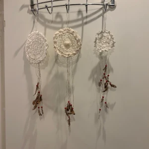 Handgjorda drömfångare med virkade detaljer - Tre handgjorda drömfångare med virkade cirklar i vitt och beige. Varje drömfångare har hängande dekorationer med fjädrar och röda pärlor. Perfekt som boho-inspirerad inredningsdetalj.