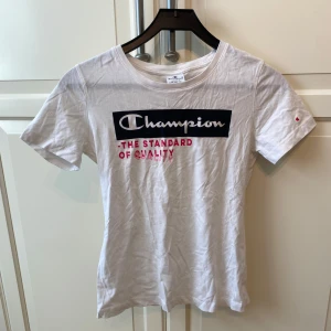 Vit t-shirt från Champion med tryck - Vit figur sydd t-shirt från Champion med svart och rött tryck på bröstet där det står 'Champion - The Standard of Quality'. Klassisk rund hals och korta ärmar med liten Champion-logga på ärmen. Tillverkad i mjuk bomull. I strl S