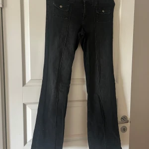 Svarta bootcut jeans med fickdetaljer - Svarta jeans med bootcut-passform och två fickor med lock framtill. Snygga kontrastsömmar och klassisk design som passar till många olika stilar. Perfekta för dig som gillar en lite vidare benslut.