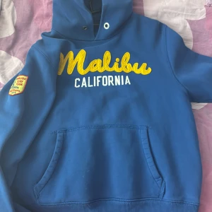 Blå hoodie med Malibu-tryck - Blå hoodie med gul och vit text 'Malibu California' på bröstet och patch på ärmen. Stor känguruficka framtill och huva med dragsko. Perfekt för en avslappnad stil.