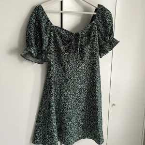 Mörkrön blommig klänning från SHEIN - Supersöt grön klänning från SHEIN med vitt blommönster och puffiga korta ärmar. Klänningen har en söt knytning framtill, dragkedja på baksidan och en luftig passform, perfekt för varma dagar. Materialet känns lätt och bekvämt. 